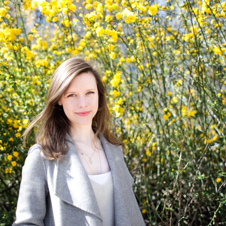 Eine junge Frau mit langen Haaren und einem lächelnden Gesichtsausdruck steht vor einem Hintergrund von leuchtend gelben Blumen.  | © privat