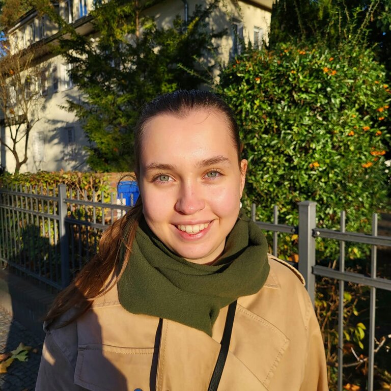 Porträtbild der Autorin Linda Pötzsch, die braune Haare hat und einen grünen Schal zusammen mit einem beigen Trenchcoat trägt. Sie lächelt dabei direkt in die Kamera. | © Linda Pötzsch
