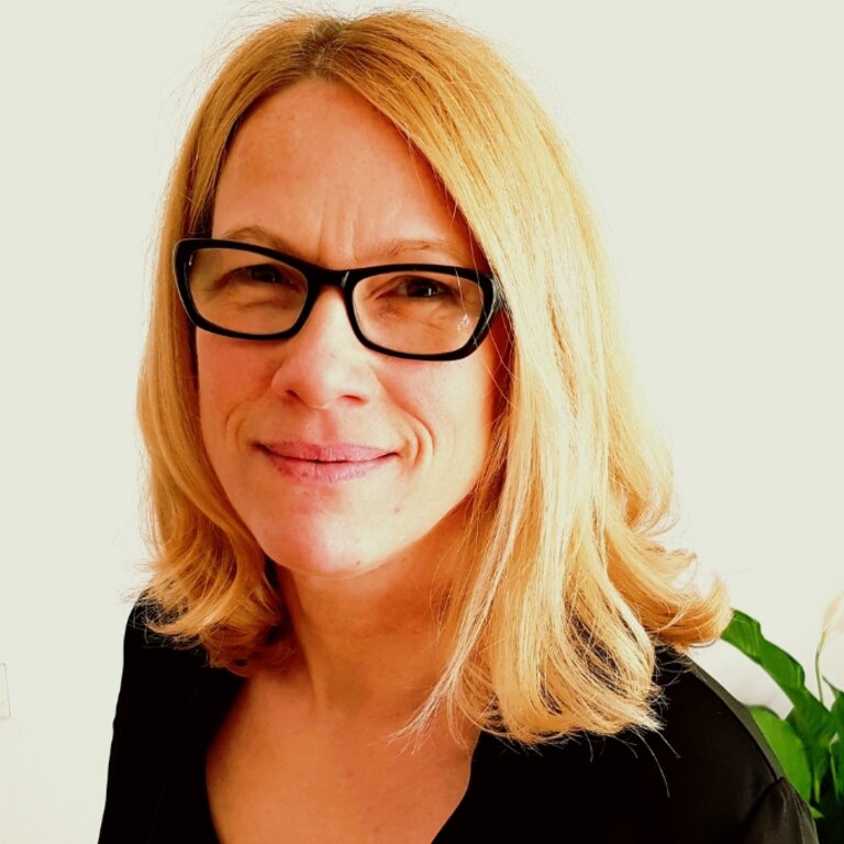 Das Bild zeigt eine lächelnde Frau mit blondem, schulterlangem Haar, die eine schwarze Brille trägt. Sie blickt direkt in die Kamera und hat einen freundlichen Gesichtsausdruck. Sie ist in einem Innenraum vor einem Hintergrund, der teilweise eine weiße Wand und eine grüne Pflanze zeigt, was eine häusliche oder büroähnliche Umgebung suggeriert. Die Frau trägt eine schwarze Kleidung, was auf einen formellen oder beruflichen Kontext hindeuten könnte. | © privat
