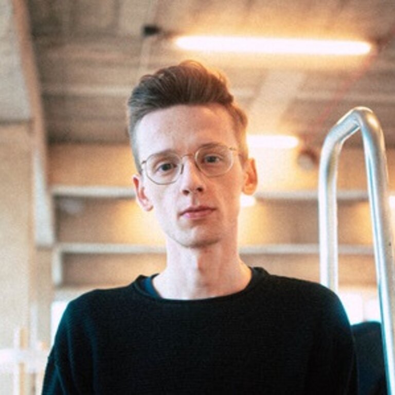 Ein junger Mann mit Brille und ordentlichem Haarschnitt trägt einen schwarzen Pullover und steht in einem Innenraum, der wie ein modernes Büro oder Café wirkt, mit unscharfem Hintergrund, der Bewegung und Geschäftigkeit suggeriert. | © privat