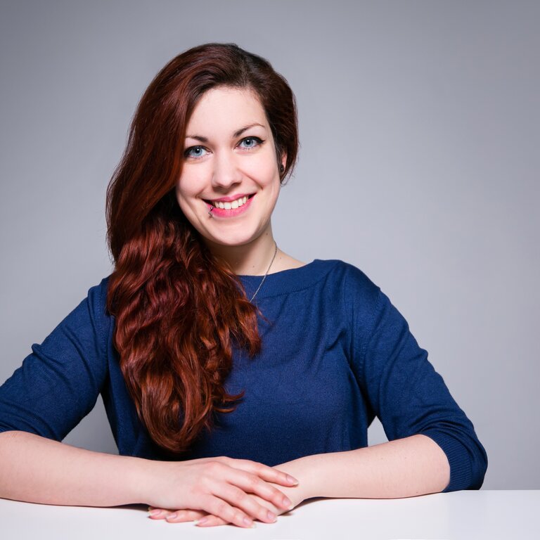 Eine Frau mit langen, welligen kastanienbraunen Haaren lächelt in die Kamera, während sie entspannt an einem Tisch sitzt und einen dunkelblauen Pullover trägt, vor einem grauen Hintergrund. | © privat