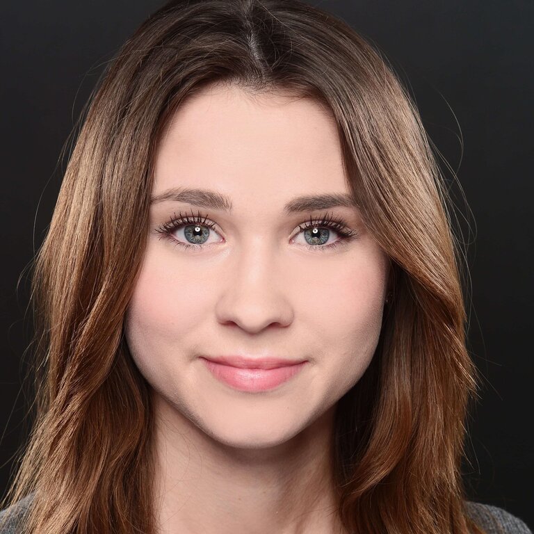 Porträt einer jungen Frau mit mittellangen brünetten Haaren und klaren blauen Augen, die direkt in die Kamera blickt und ein subtiles Lächeln zeigt, vor einem dunklen Hintergrund. | © privat