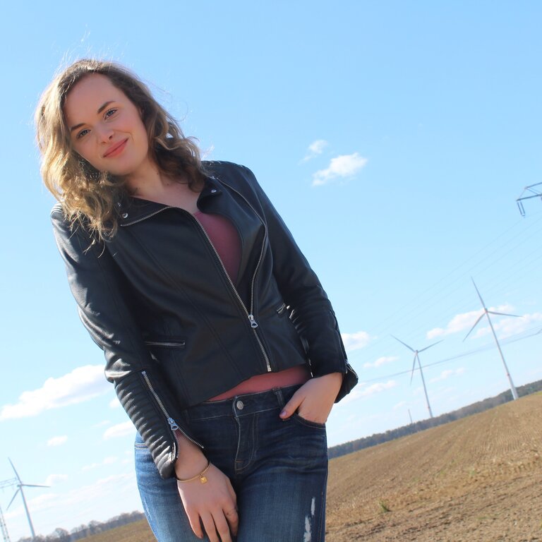 Das Bild zeigt eine junge Frau, die draußen steht und selbstbewusst in die Kamera lächelt. Sie trägt eine schwarze Lederjacke, ein dunkles Oberteil und Jeans. Im Hintergrund ist eine ländliche Landschaft mit einem klaren blauen Himmel, Windrädern und einem Strommast zu sehen. Die Pose der Frau ist entspannt und lässig, und sie scheint die frische Luft und die Umgebung zu genießen. | © privat