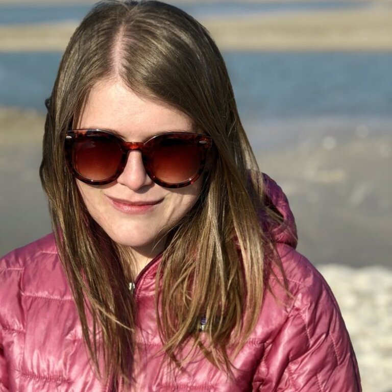 Das Bild zeigt eine Frau mit blonden Haaren und Sonnenbrille, die in die Kamera lächelt. Sie trägt eine pinke Jacke und scheint sich an einem Strand oder einer Küstenregion zu befinden, erkennbar an dem Sand und Wasser im Hintergrund. Die Sonne scheint auf ihr Gesicht, was ihr einen fröhlichen Ausdruck verleiht. | © privat
