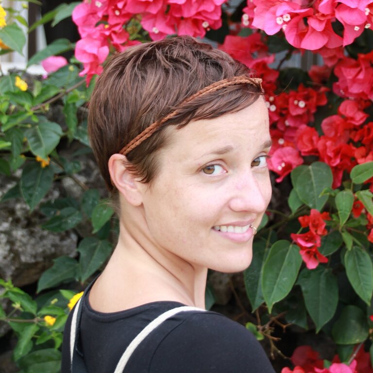 Eine Frau mit kurzen Haaren steht vor einer blumigen Kulisse und lächelt in die Kamera.  | © privat