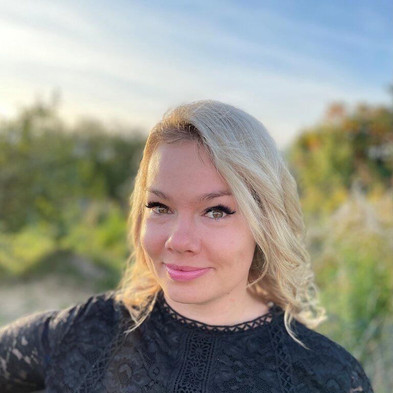 Blonde, junge Frau lächelt in die Kamera. Sie trägt ein schwarzes Oberteil und steht im Freien. Im Hintergrund erkennt man verschwommen Bäume. Madeleine Liedke ist Online-Redakteurin bei BILD der FRAU.  | © Anne Zeidler 