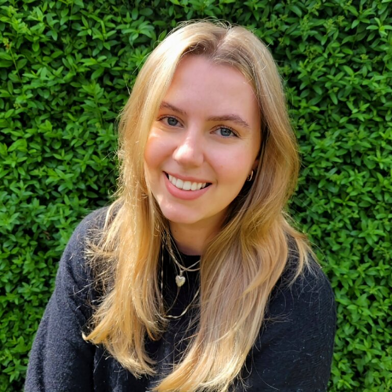 Das Bild zeigt eine Frau mit blonden, langen Haaren und blauen Augen, die freundlich in die Kamera lächelt. Sie sitzt seitlich vor einem grünen Hintergrund und trägt ein schwarzes Oberteil.  | © privat