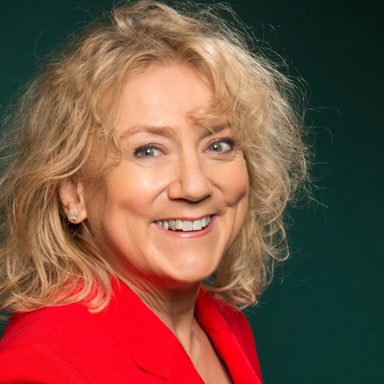 Das Bild zeigt eine strahlende Frau mit lockigen blonden Haaren, die in die Kamera lächelt. Sie trägt einen leuchtend roten Blazer, der einen fröhlichen und energischen Eindruck macht. | © batchfield