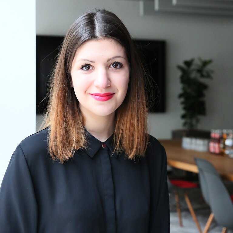 Eine junge Frau mit schulterlangem braunem Haar und einem schwarzen Hemd lächelt in die Kamera, vor dem Hintergrund eines modernen Bürointerieurs mit Pflanzen und Designermöbeln | © Mandy Buchholz