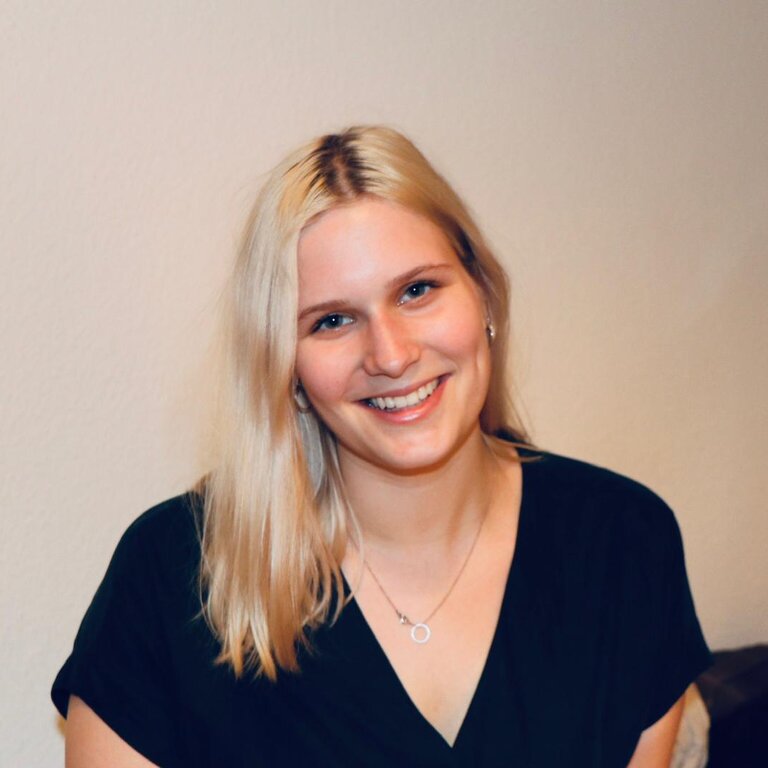 Das Bild zeigt eine junge Frau mit blonden Haaren, die in die Kamera lächelt. Sie trägt ein schwarzes Oberteil und eine Kette mit einem kleinen Anhänger. Ihr freundliches Lächeln und ihre offene Körperhaltung vermitteln eine positive und herzliche Ausstrahlung. Der Hintergrund ist neutral und unscharf, was den Fokus auf die Frau richtet. | © privat