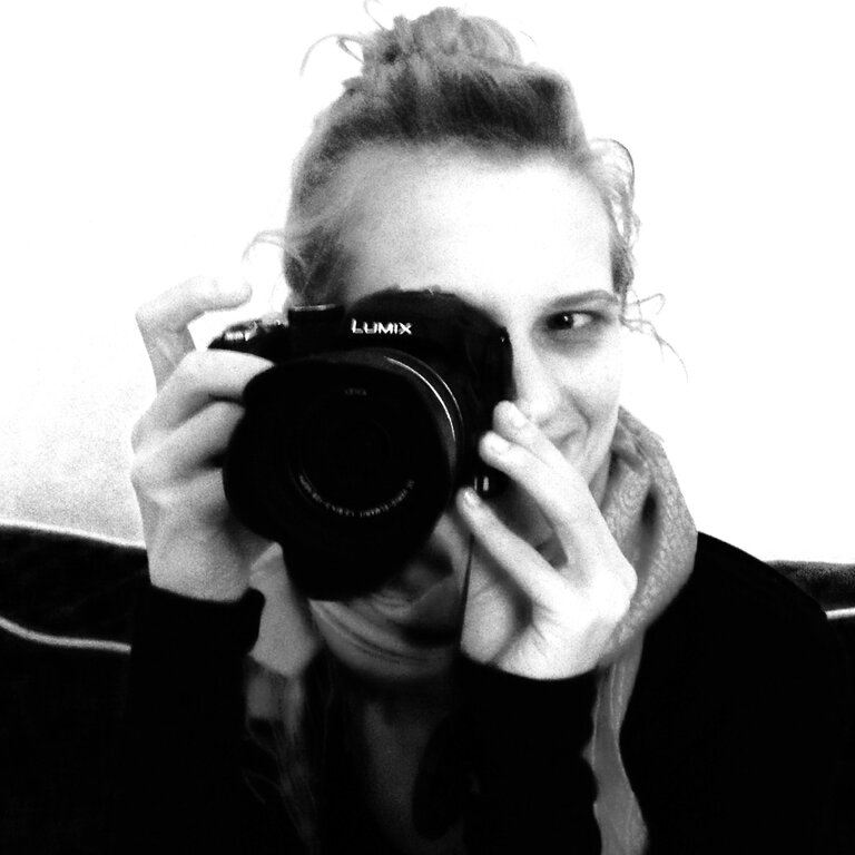 Schwarz-weiß Foto einer lächelnden Person, die eine Lumix Kamera vor ihr Gesicht hält, um ein Foto zu machen, wobei nur ein Auge sichtbar ist, was eine lebendige und kreative Atmosphäre schafft. | © privat