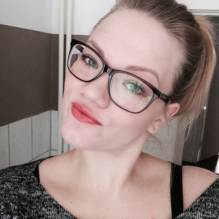 Selbstporträt einer Frau mit zurückgebundenen blonden Haaren und großer Brille, die leuchtend roten Lippenstift trägt und direkt in die Kamera blickt. | © privat