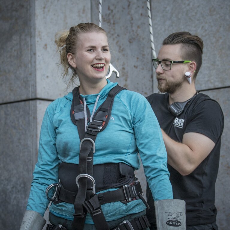 Eine lächelnde Frau in Kletterausrüstung und blauer Sportkleidung steht neben einem Mann mit Brille und Kopfhörern. Sie scheint sich auf eine Kletteraktivität vorzubereiten, während der Mann im Hintergrund steht und ein Sicherungsgerät hält, beide vor einer grauen Betonwand | © Pascal Rohe