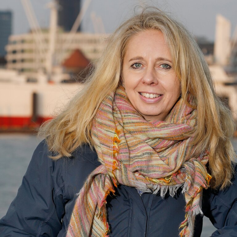 Eine lächelnde blonde Frau, bekleidet mit einem dunklen Wintermantel und einem farbenfrohen Schal, steht vor einer städtischen Flusskulisse mit modernen Gebäuden und Kränen im Hintergrund, was auf eine lebendige Hafenatmosphäre hindeutet. | © Sybill Schneider