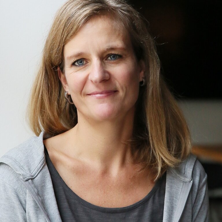 Das Bild zeigt eine Frau mit freundlichem Lächeln, die direkt in die Kamera blickt. Sie hat mittellanges blondes Haar, blaue Augen und trägt ein graues T-Shirt mit einer offenen grauen Jacke. Der Hintergrund ist schlicht und unauffällig, was den Fokus auf die Frau lenkt. Ihr Ausdruck wirkt offen und zugänglich. | © BILD der FRAU