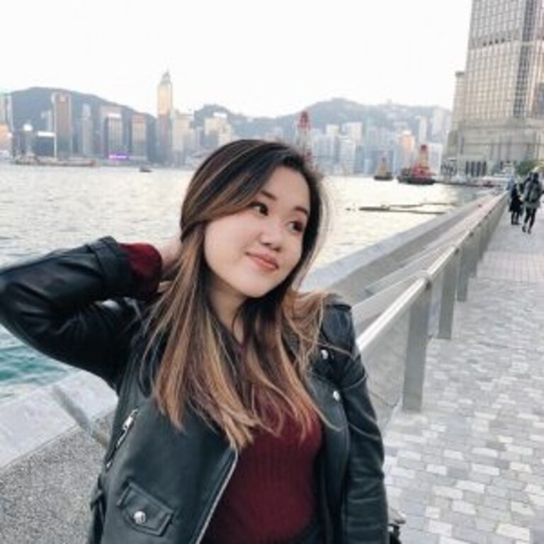 Eine junge Frau mit langem, welligem Haar und einer schwarzen Lederjacke steht lächelnd am Ufer einer Hafenpromenade. Sie hat eine Hand leicht in ihrem Haar und blickt träumerisch zur Seite. Im Hintergrund ist eine Stadtlandschaft mit Hochhäusern und der Silhouette eines bewölkten Himmels über einer Wasserfläche zu erkennen. | © privat