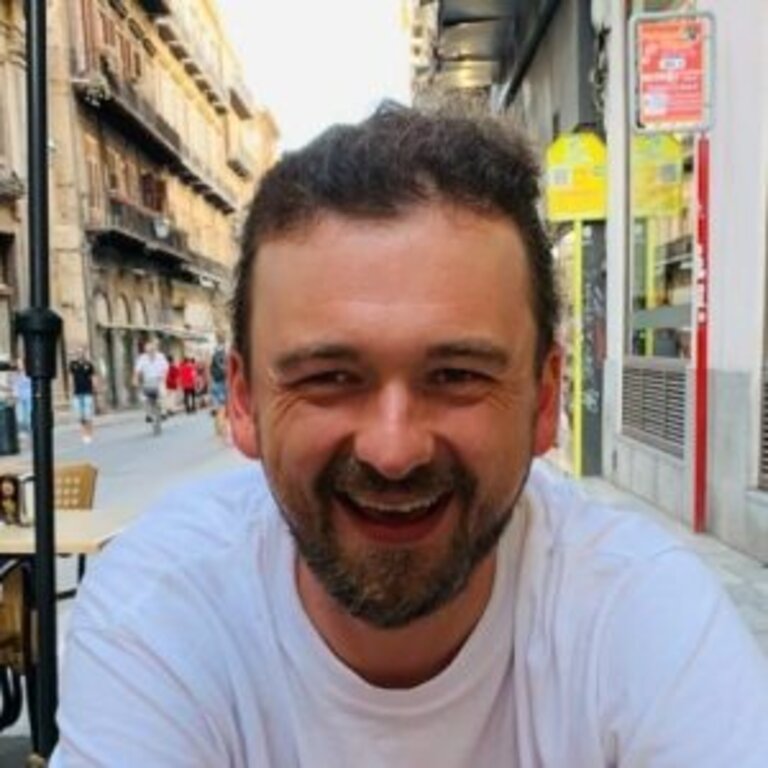 Das Bild zeigt einen lächelnden Mann mit kurzen dunklen Haaren und Bart. Er trägt ein weißes T-Shirt und sitzt im Freien, wie es scheint, in einem Straßencafé. Im Hintergrund ist eine belebte Straße mit historischen Gebäuden zu erkennen. | © privat