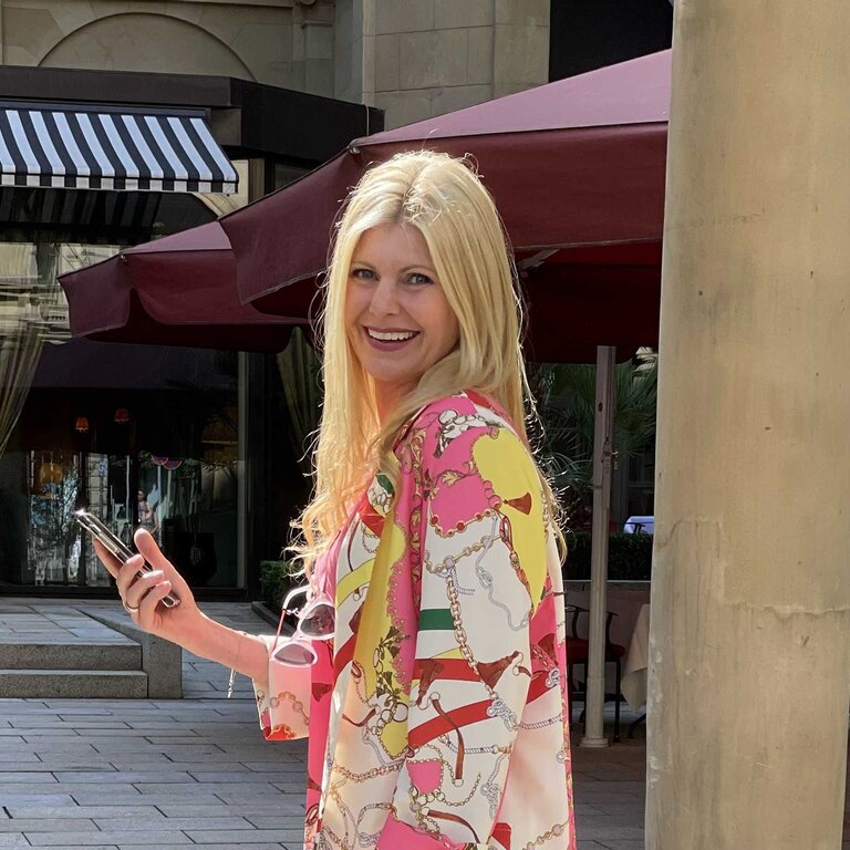 Das Bild zeigt eine Frau mit langen blonden Haaren und einem strahlenden Lächeln, die in einer städtischen Umgebung steht. Sie trägt ein farbenfrohes Outfit mit einem Muster, das an Ketten und Riemen erinnert, und strahlt eine fröhliche und entspannte Stimmung aus. | © privat