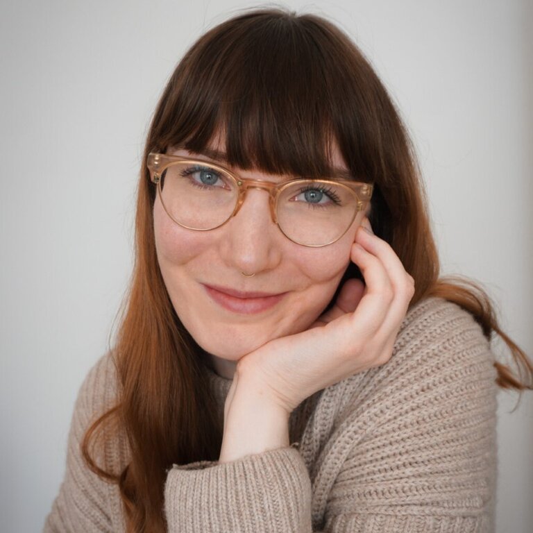 Porträt einer lächelnden Frau mit kinnlangem, glattem braunem Haar und Pony, die hellbraune, runde Brillengestelle trägt. Sie stützt ihre Wange auf ihre Hand und blickt direkt in die Kamera mit einem freundlichen Ausdruck. Sie trägt einen gemütlichen Strickpullover in einem passenden Braunton, was auf eine bequeme, zwanglose Umgebung hindeutet. Ihr dezentes Make-up und ihr natürliches Aussehen vermitteln ein Gefühl von Zugänglichkeit und Wärme. | © privat