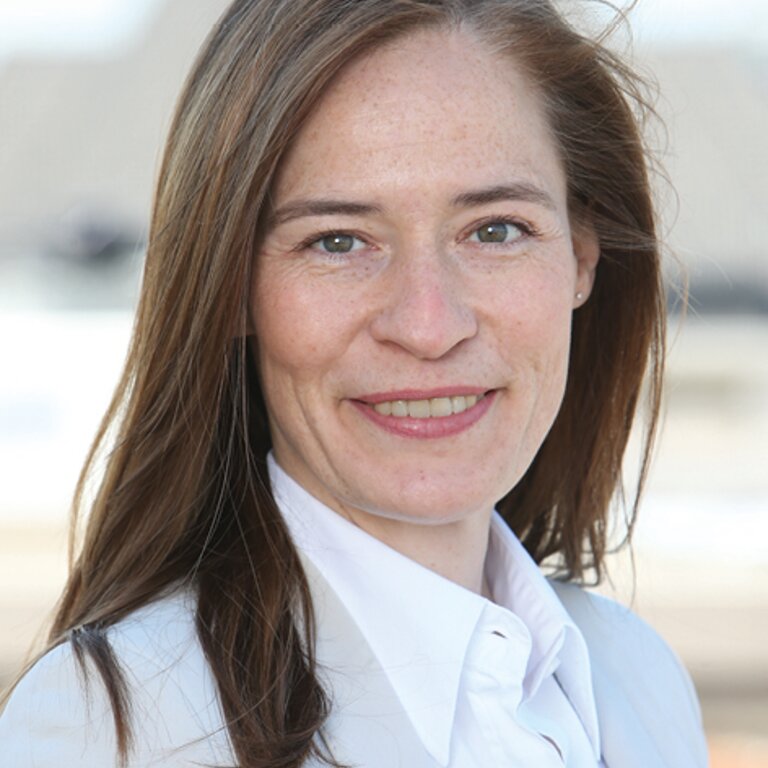 Eine Frau mit langen braunen Haaren und einem freundlichen Lächeln trägt eine weiße Bluse und posiert für ein professionelles Porträtfoto. | © privat