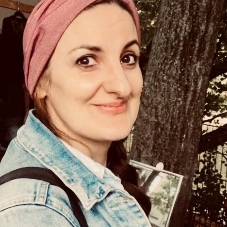 Die Frau auf dem Foto trägt ein rosa Kopftuch und eine Jeansjacke. Sie hat dunkles, zur Seite geflochtenes Haar und ein freundliches Lächeln. Der Hintergrund ist unscharf, aber es scheint, als befände sie sich im Freien, möglicherweise in einem Park, da Bäume zu sehen sind.  | © privat
