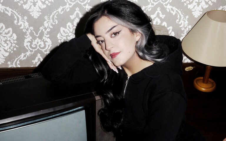 Parshad Esmaeili sitzt auf einem Teppich neben einem Fernseher und schaut nachdenklich in die Kamera.  | © Lottermann and Fuentes / Droemer Knaur