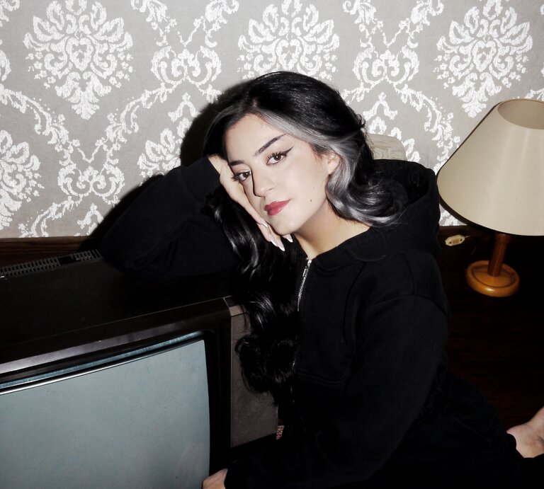 Parshad Esmaeili sitzt auf einem Teppich neben einem Fernseher und schaut nachdenklich in die Kamera.  | © Lottermann and Fuentes / Droemer Knaur