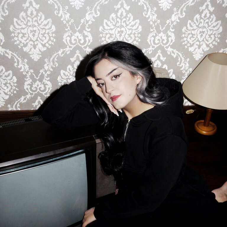 Parshad Esmaeili sitzt auf einem Teppich neben einem Fernseher und schaut nachdenklich in die Kamera.  | © Lottermann and Fuentes / Droemer Knaur