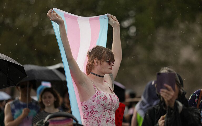 Eine Person steht in einem ärmellosen pink-weißen Top inmitten einer Menge im Regen und hält mit beiden Armen eine Trans-Pride Flagge über ihren Kopf | © Bryan Dozier | getty images