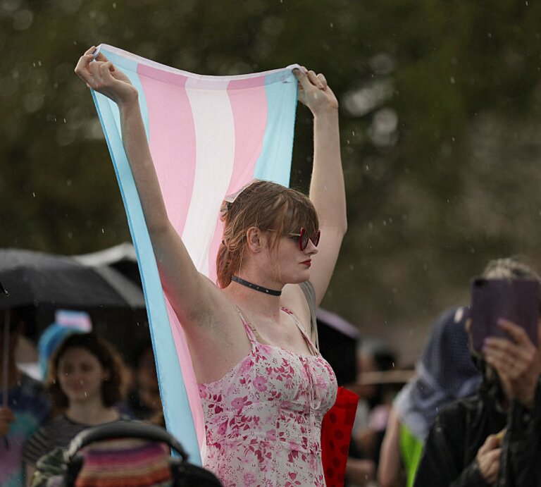 Eine Person steht in einem ärmellosen pink-weißen Top inmitten einer Menge im Regen und hält mit beiden Armen eine Trans-Pride Flagge über ihren Kopf | © Bryan Dozier | getty images