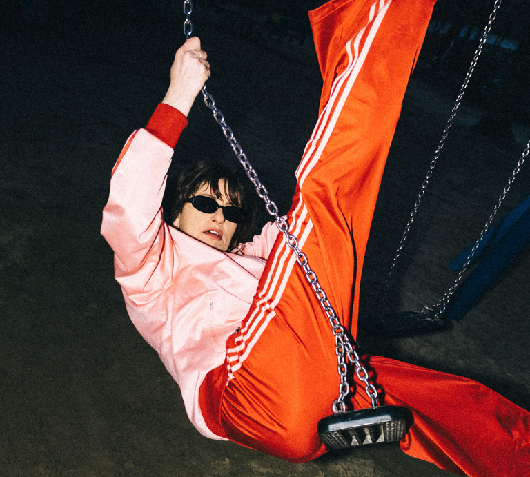 Die Künstlerin Mola sitzt auf einer Schaukel und trägt eine orangefarbene Sporthose sowie eine pinke Jacke. | © André Beiler