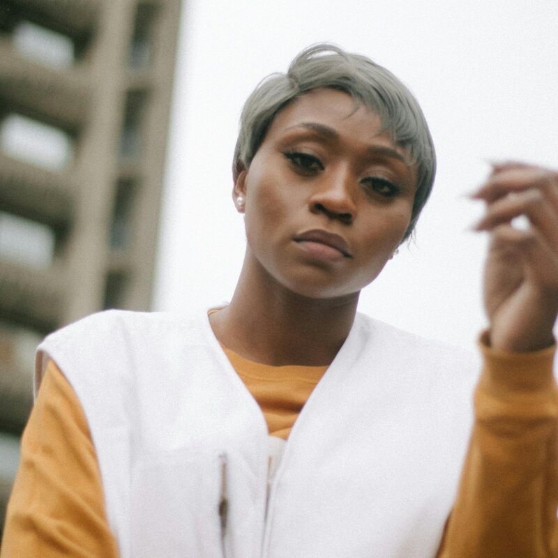 Im Hintergrund ist ein Hochhaus zu sehen, im Vordergrund eine ernst schauende Frau.  | © Ezekixl Akinnewu | Pexels 