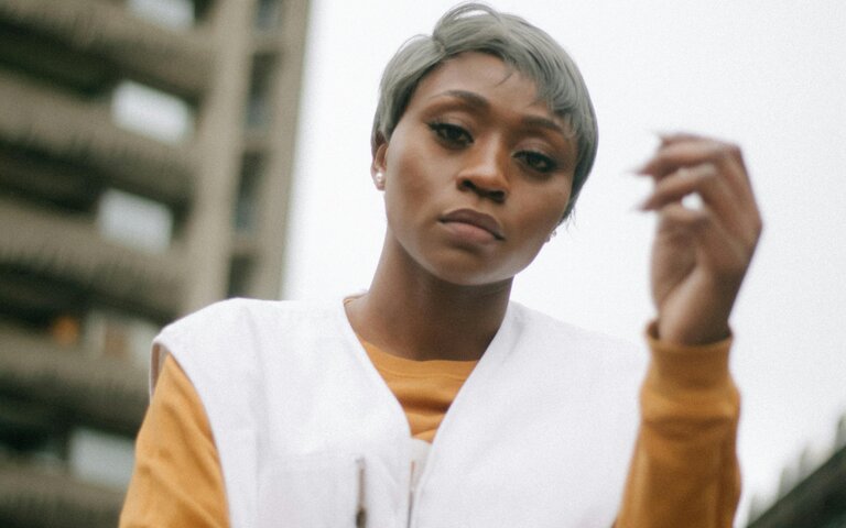 Im Hintergrund ist ein Hochhaus zu sehen, im Vordergrund eine ernst schauende Frau.  | © Ezekixl Akinnewu | Pexels 