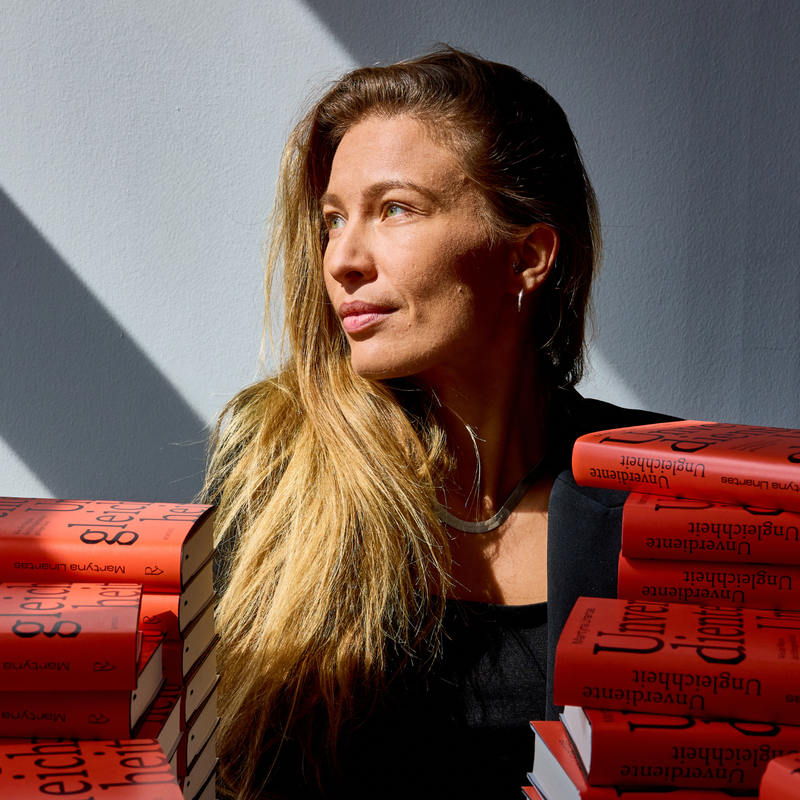 Porträt von Dr. Martyna Linartas, die hinter einem Stapel ihrer Bücher sitzt.  | © Norman Konrad | Dr. Martyna Linartas beschreibt in ihrem Buch, wie der Weg aus der sogenannten Erbengesellschaft gelingen kann. 