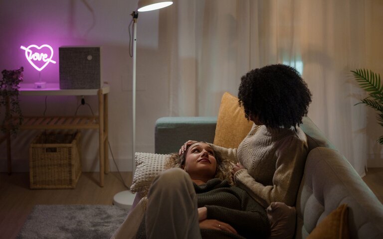 Zwei Personen sitzen auf einem Sofa bei leicht gedimmtem Licht. Die eine Person streichelt der anderen Person sanft über den Kopf. | © Getty Images/Ivan Rodriguez Alba
