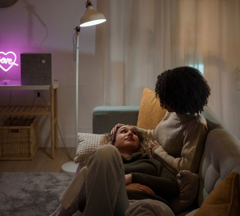 Zwei Personen sitzen auf einem Sofa bei leicht gedimmtem Licht. Die eine Person streichelt der anderen Person sanft über den Kopf. | © Getty Images/Ivan Rodriguez Alba