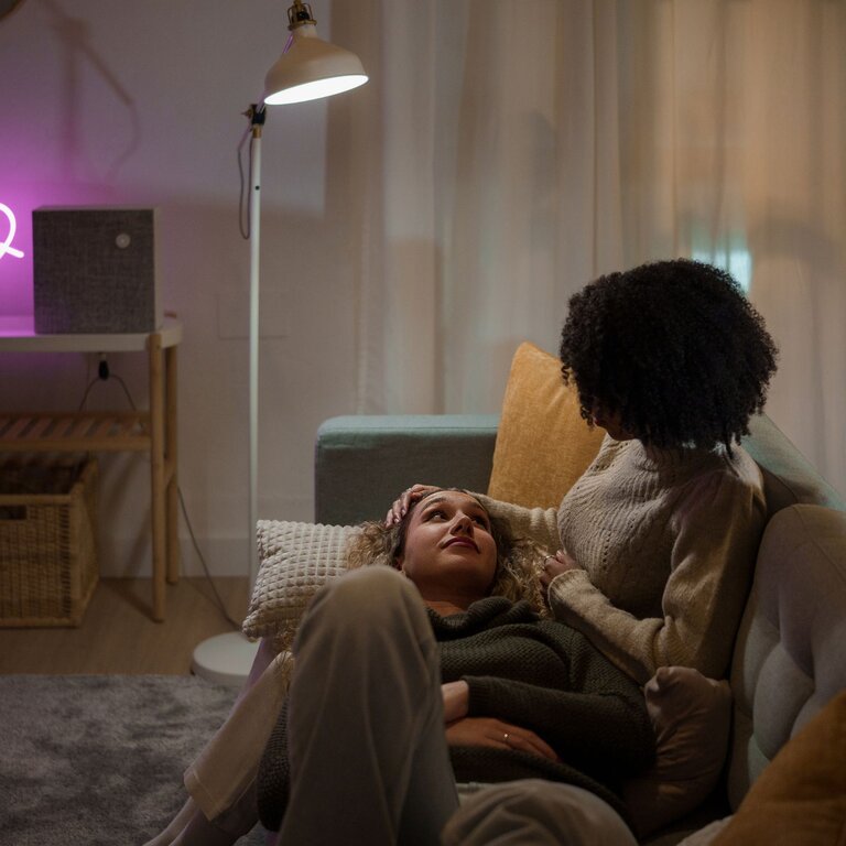 Zwei Personen sitzen auf einem Sofa bei leicht gedimmtem Licht. Die eine Person streichelt der anderen Person sanft über den Kopf. | © Getty Images/Ivan Rodriguez Alba