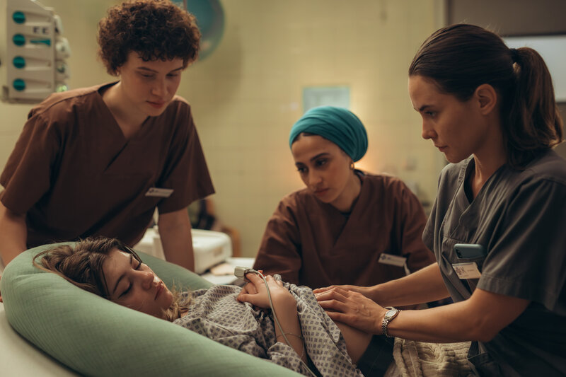 "PUSH - Geheimnisse": Lou (Lisa Moell) liegt in der Klinik. Greta (Lydia Amasko) und Meryem (Tua El-Fawwal) stehen daneben während Dr. Mohn (Katia Fellin) Lou untersucht. | © ZDF | Andrea Hansen: Lou bringt ihr Kind anonym zur Welt. Greta (Lydia Amasko, l.) und Meryem sind für die junge Frau da. Das Kind wird direkt nach der Geburt zur Adoption freigegeben. 