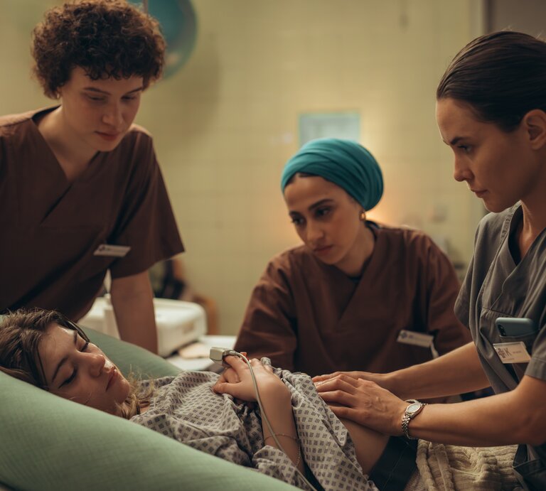 "PUSH - Geheimnisse": Lou (Lisa Moell) liegt in der Klinik. Greta (Lydia Amasko) und Meryem (Tua El-Fawwal) stehen daneben während Dr. Mohn (Katia Fellin) Lou untersucht. | © ZDF | Andrea Hansen: Lou bringt ihr Kind anonym zur Welt. Greta (Lydia Amasko, l.) und Meryem sind für die junge Frau da. Das Kind wird direkt nach der Geburt zur Adoption freigegeben. 