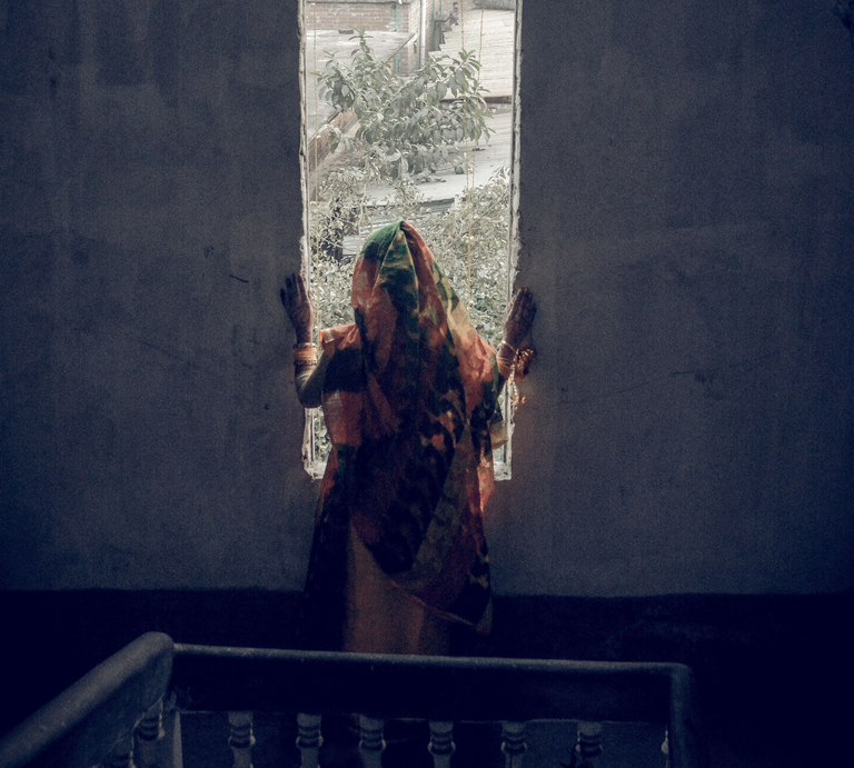 Eine Frau in traditioneller Kleidung steht in einem unheimlichen, verlassenen Treppenhaus, eingerahmt von einem Fenster. | ©  Sadman Chowdhury | Pixels