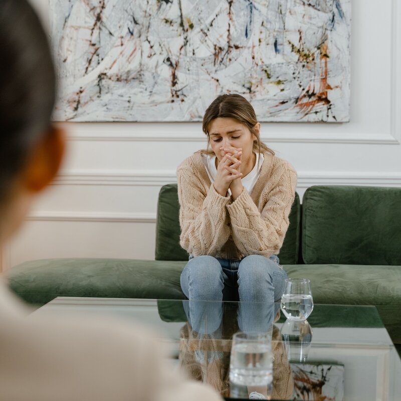 Junge Frau sitzt bei der Therapeutin, wirkt verzweifelt. Helles Sofa. Frau hat beigebraune Jacke. Kopf in die Hände gelegt.  | © Mart Production | Pexels