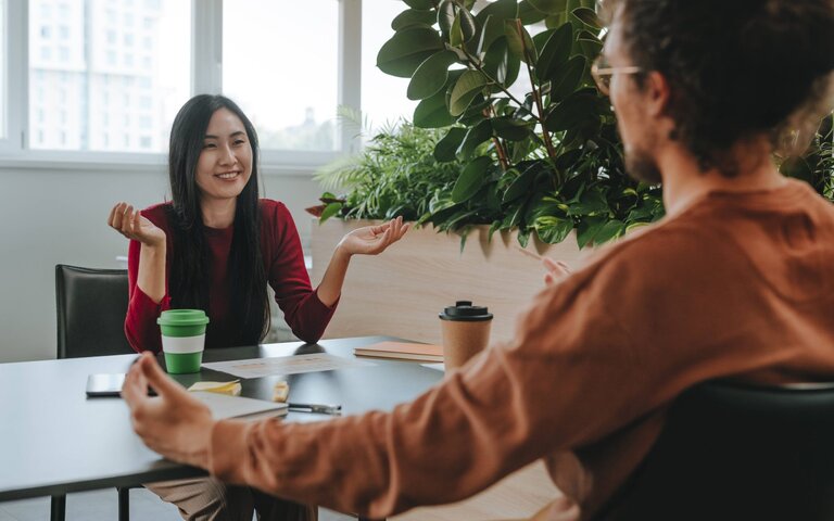 Zwei Personen sitzen sich an einem Arbeitstisch gegenüber. | © IMAGO / Westend61