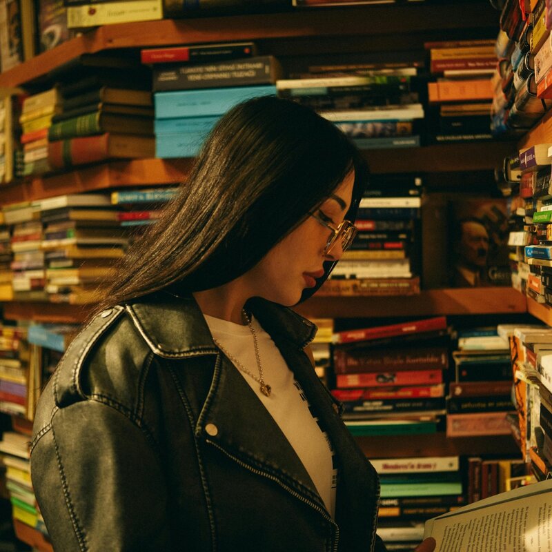 Junge Frau steht vor einem riesigen Bücherregal und liest eines der bunten Bücher. Sie trägt eine Brille und langes schwarzes Haar. | © Foto: Göksu Taymaz | Pexels