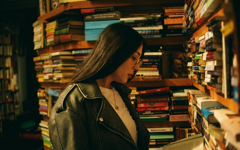 Junge Frau steht vor einem riesigen Bücherregal und liest eines der bunten Bücher. Sie trägt eine Brille und langes schwarzes Haar. | © Foto: Göksu Taymaz | Pexels