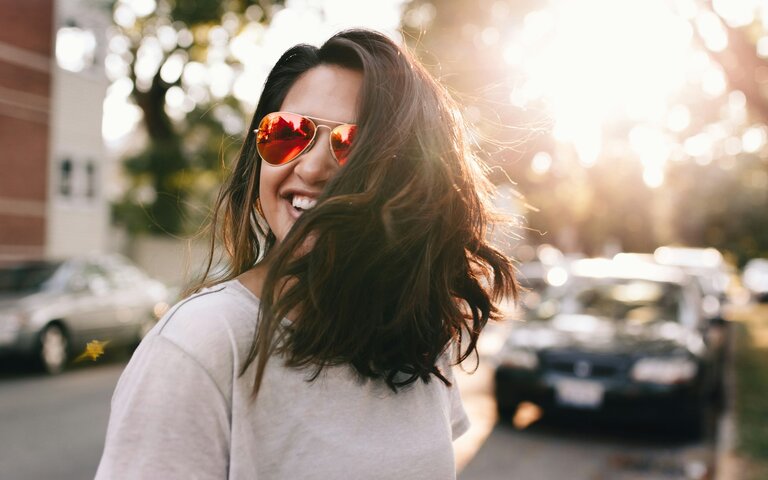 Frau mit roter Sonnenbrille und langem dunklem Haar lacht auf der Straße, sie wirkt glücklich. | © Foto: Matthew Hamilton | Unsplash 