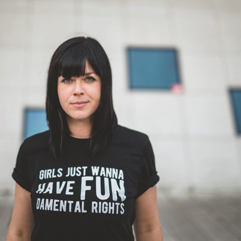 Mareike Fallwickl, Autorin des Buches „Die Wut, die bleibt“ steht im öffentlichen Raum vor einem Haus mit wenigen dunklen Spiegelfenstern. Auf ihrem T-Shirt steht der Schriftzug: Girls just wanna have fun. Demental Rights. : | © Foto: Gyöngyi Tasi