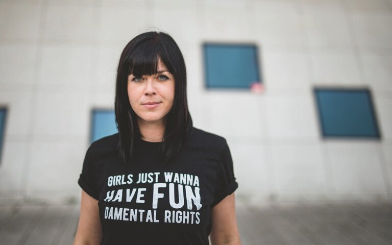 Mareike Fallwickl, Autorin des Buches „Die Wut, die bleibt“ steht im öffentlichen Raum vor einem Haus mit wenigen dunklen Spiegelfenstern. Auf ihrem T-Shirt steht der Schriftzug: Girls just wanna have fun. Demental Rights. : | © Foto: Gyöngyi Tasi