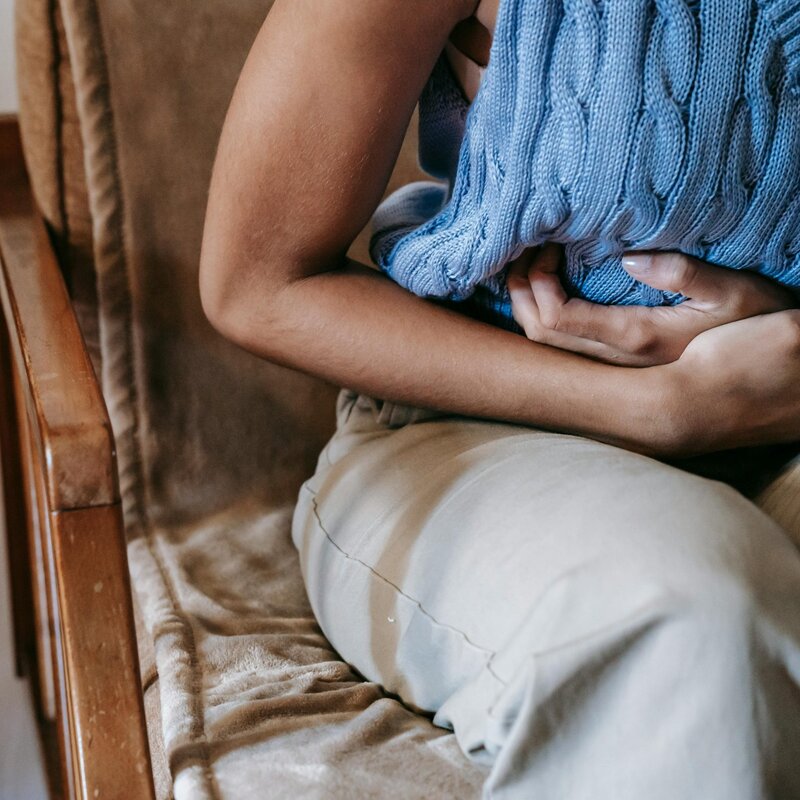 Frau hält sich den Bauch, trägt blaues armfreies gestricktes Shirt und eine beige Hose, Kopf ist nicht zu erkennen. | © Foto: Sora Shimazaki | Pexels