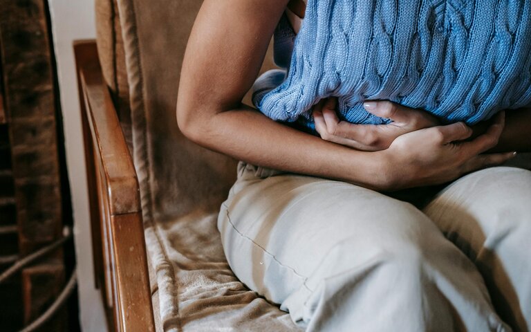 Frau hält sich den Bauch, trägt blaues armfreies gestricktes Shirt und eine beige Hose, Kopf ist nicht zu erkennen. | © Foto: Sora Shimazaki | Pexels