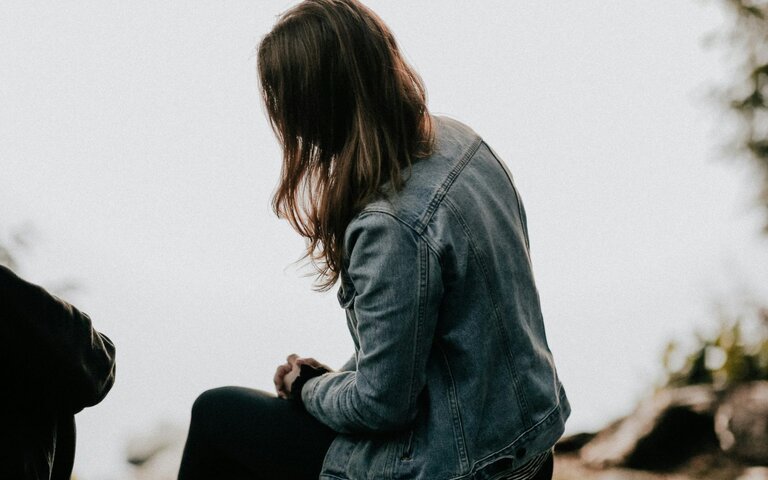 Frau sitzt am Wasser, Jeansjacke, lange braune Haare. Hintergrund grau. | © Foto: Priscilla du Preez | Unsplash